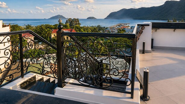 Un balcon doté d’une balustrade en fer forgé, offrant une vue sur la côte, avec des montagnes et une étendue d’eau.