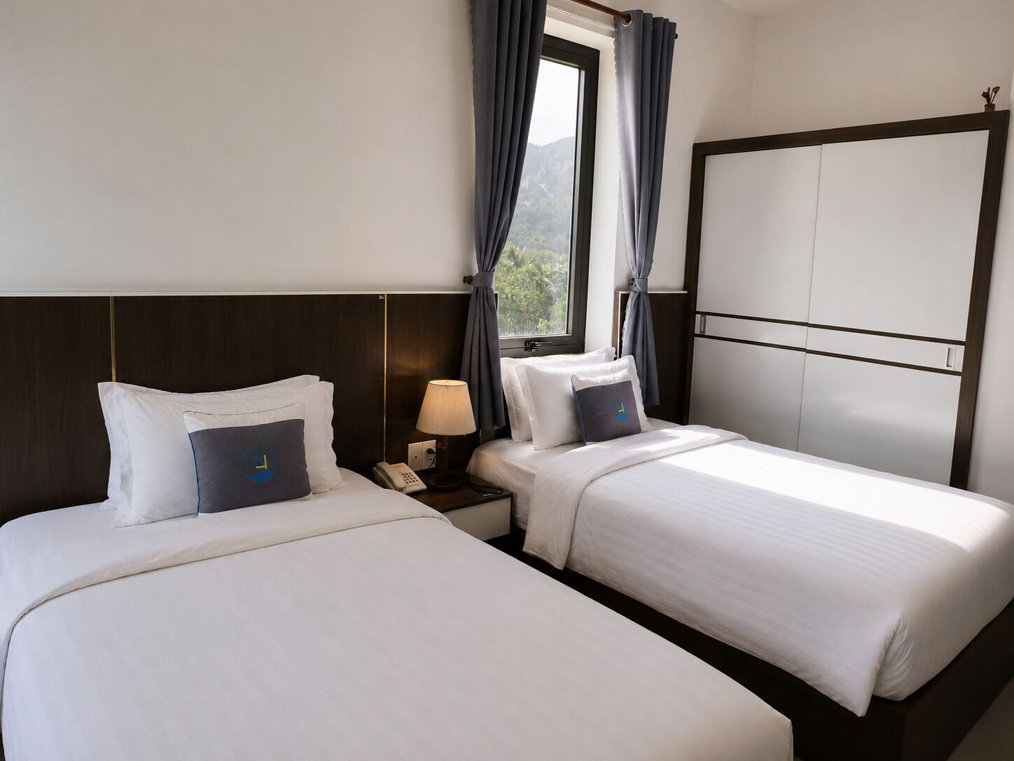 Una habitación de hotel con dos camas, una mesita de noche con lámpara, teléfono y armario.