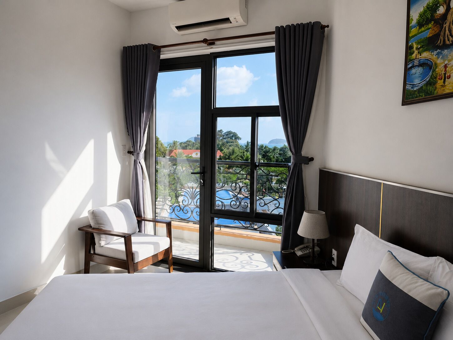 Habitación de hotel con una cama, una silla, una mesita de noche con lámpara, y vistas a una piscina y edificios a través de un ventanal grande.