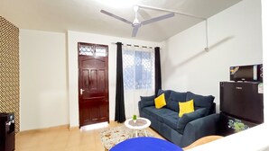A living room with a ceiling fan, a dark blue sofa with yellow cushions, a small white table, and a black refrigerator. - Sunset Suites Mtwapa (Mtwapa)