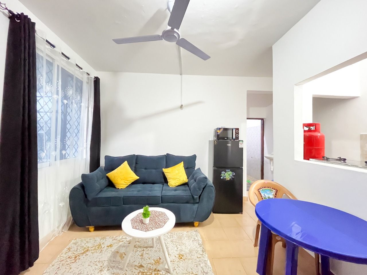 A living room with a dark blue sofa, yellow cushions, a small white table with a plant, a ceiling fan, a black refrigerator, a blue table, and a red gas cylinder.
