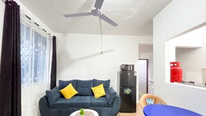 A living room with a dark blue sofa, yellow cushions, a small white table with a plant, a ceiling fan, a black refrigerator, a blue table, and a red gas cylinder. - Sunset Suites Mtwapa (Mtwapa)