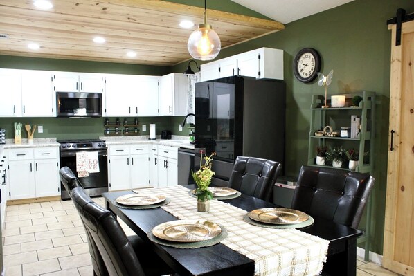 A modern kitchen with white cabinets, a black refrigerator, and a dining area with a black table set for four. - Coastal House Near FLETC, St Simon's & Jekyll Island (Brunswick)
