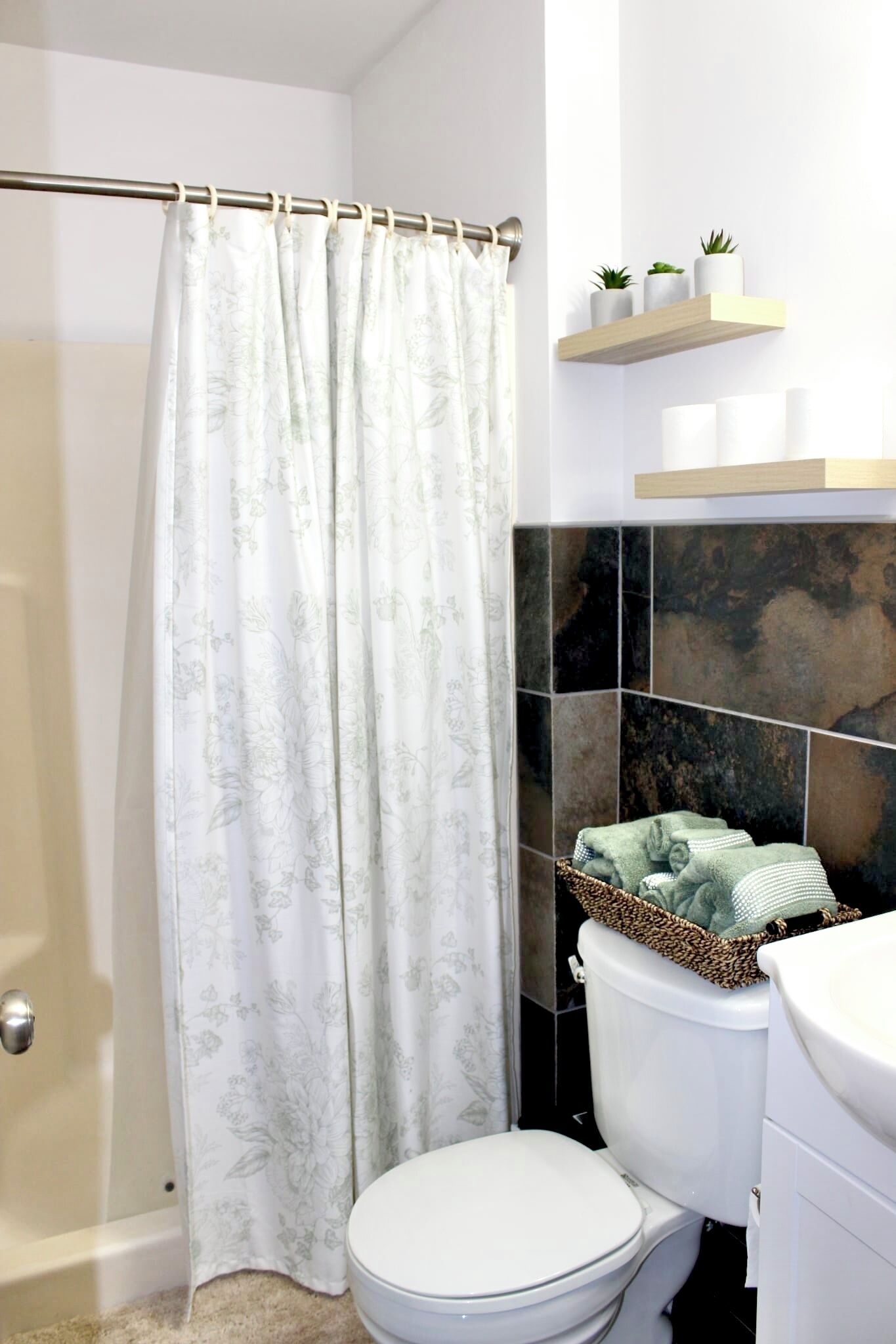 A bathroom with a shower curtain, a toilet, a sink, and a shelf with plants.