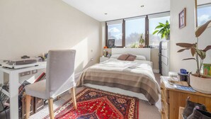 Une chambre moderne avec un grand lit, un bureau et une chaise. De nombreuses fenêtres laissent entrer la lumière naturelle et offrent une vue sur l’extérieur.