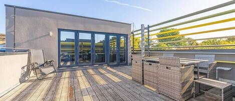 A rooftop terrace with a modern building, outdoor furniture, and a wooden deck.