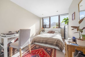 Ein modernes Schlafzimmer mit einem großen Bett, einem Schreibtisch und einem Sessel. Mehrere Fenster sorgen für natürliches Licht und einen Ausblick ins Freie.