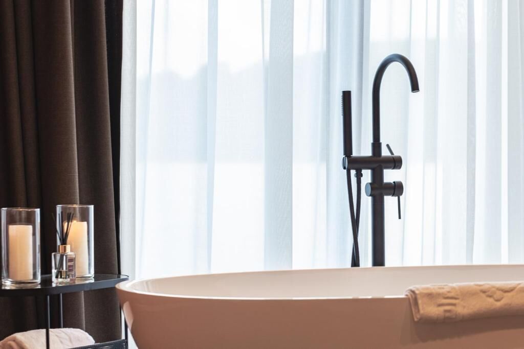 A modern bathroom with a freestanding bathtub, a black faucet, and a window with sheer curtains.
