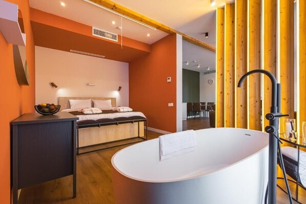 A modern bedroom with a large bed, a bedside table with a bowl of fruit, a white bathtub, and wooden paneling on the wall. - Kundzuli Apart Hotel  (Kvareli)