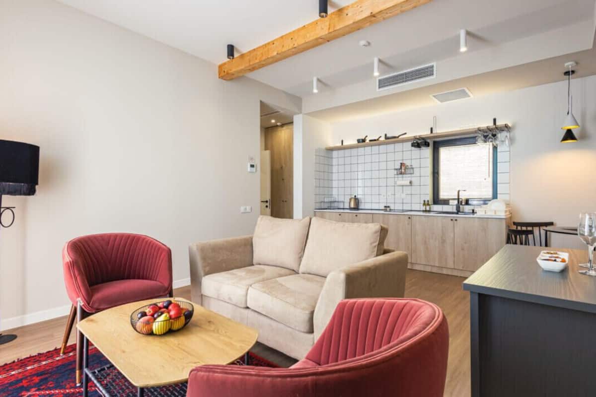A modern living room with a sofa, two armchairs, a coffee table with fruit, and a kitchen in the background.