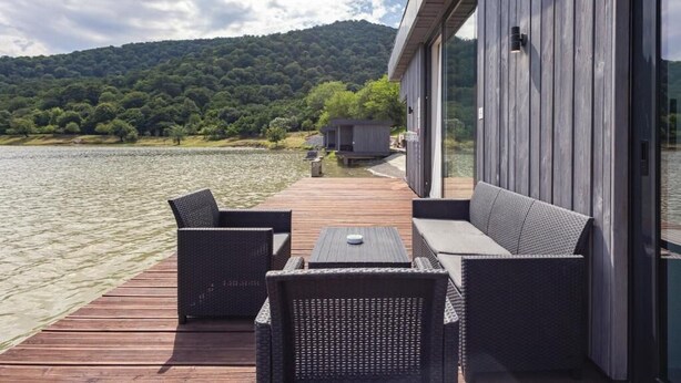 Eine Terrasse mit Korbmöbeln mit Blick auf einen See, ein Gebäude aus Holz mit großen Fenstern und ein bewaldeter Hügel im Hintergrund.