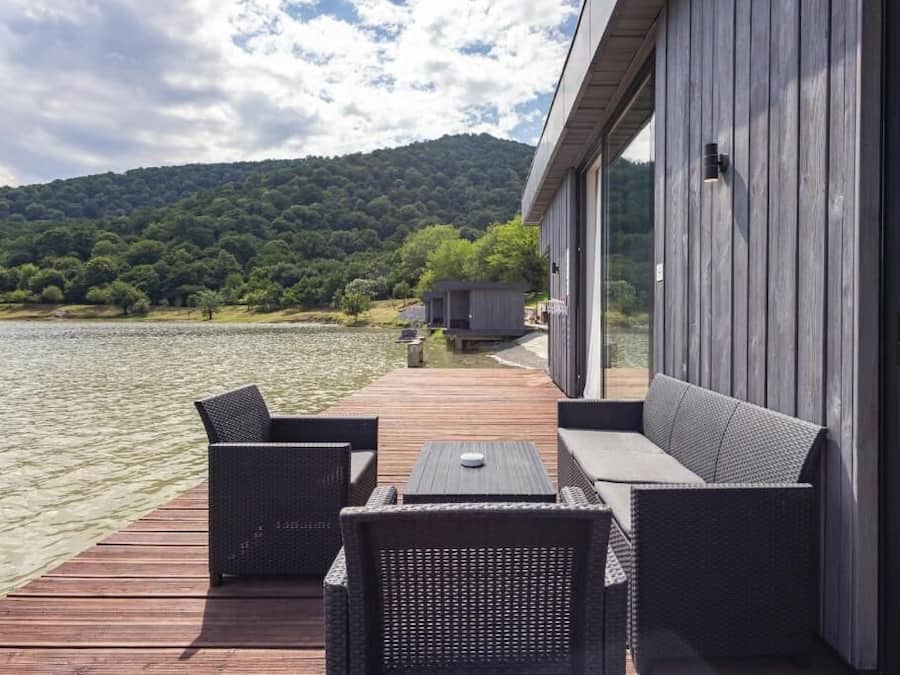 A deck with wicker furniture overlooking a lake, a wooden building with large windows, and a forested hill in the background.