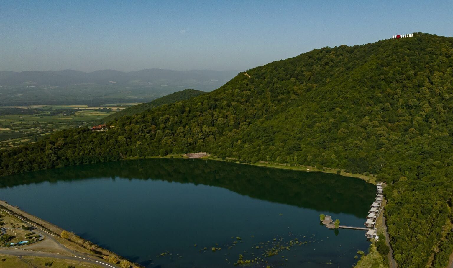 Um lago rodeado por colinas florestadas, com um pequeno cais e uma fileira de cabanas.