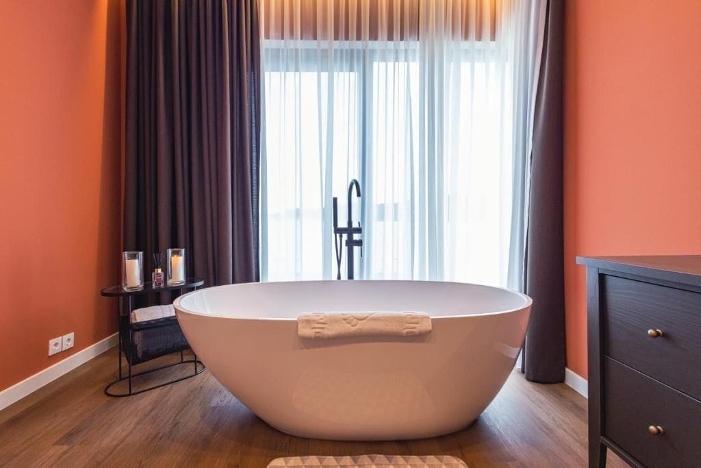 A modern bathroom with a freestanding bathtub, a vanity with drawers, and a window with sheer curtains.