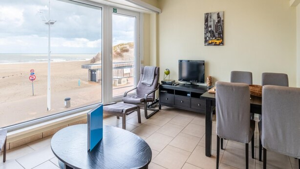 A room with a large window overlooking a beach, a television, a dining table with chairs, and a small seating area with a chair and a table. - Cosy flat with frontal sea view (Koksijde)
