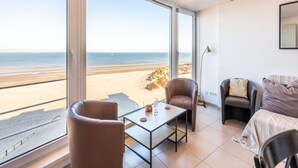 A room with a large window overlooking a beach, a table with a glass, two chairs, and a sofa. - Renovated flat with sea view (Koksijde)