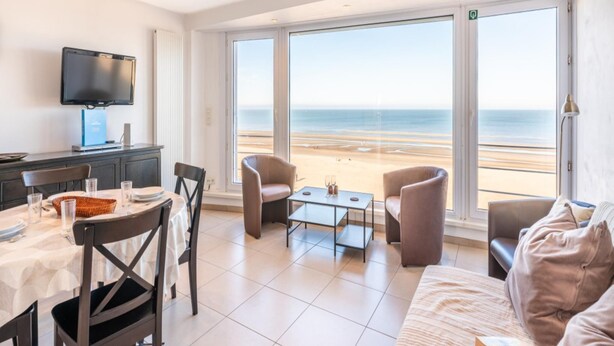 A hotel room with a large window offering a beach view, a dining area with a table and chairs, a flat-screen TV, and a sofa. - Renovated flat with sea view (Koksijde)