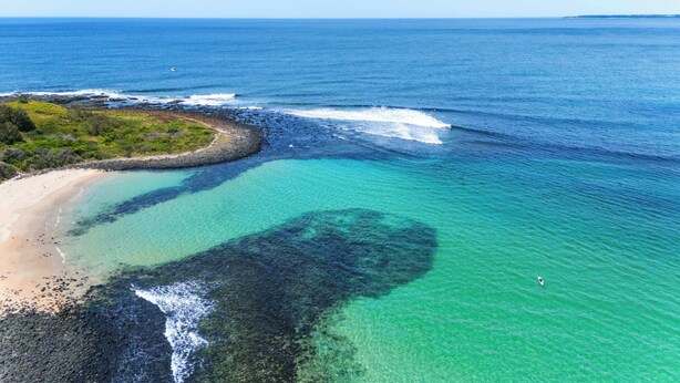 An aerial view of a coastal area with clear turquoise waters, a sandy beach, and a rocky shoreline.