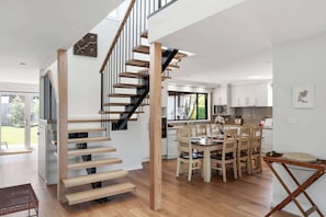 Eine moderne Küche mit weißen Schränken, ein Essbereich mit Holzstühlen sowie eine Treppe mit Holzstufen und schwarzen Geländern.