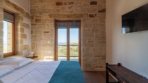 Une chambre avec un mur en pierre, un lit recouvert de draps blancs, une couverture bleue, un banc en bois et un téléviseur fixé au mur.