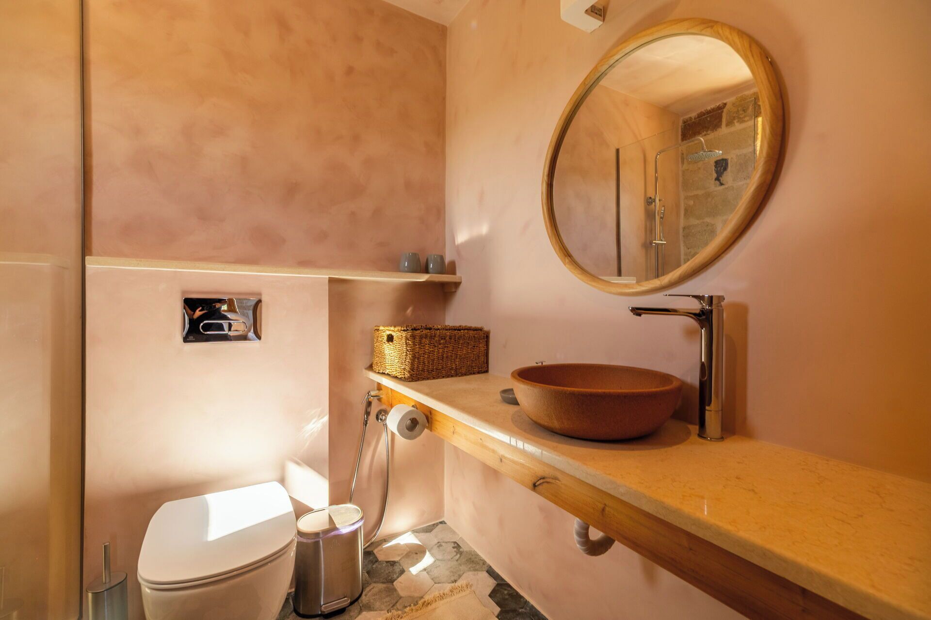 A bathroom with a wooden vanity, a round mirror, a bowl sink, a toilet, and a shower with a glass door.