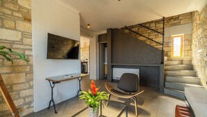A modern living room with a stone wall, a flat-screen TV, a glass coffee table, a comfortable chair, and a staircase with a metal railing. - Villa Seiri – Vacation villa in Crete (Gavalochori)