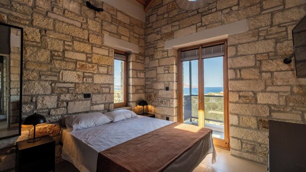 A bedroom with a stone wall, a bed, a nightstand, a lamp, and a view of the sea. - Villa Seiri – Vacation villa in Crete (Gavalochori)