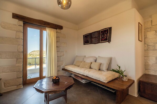 A cozy room with a stone wall, a wooden bench, a sofa, and a table with a bowl of bread. - Villa Falcos– Vacation villa in Crete (Gavalochori)