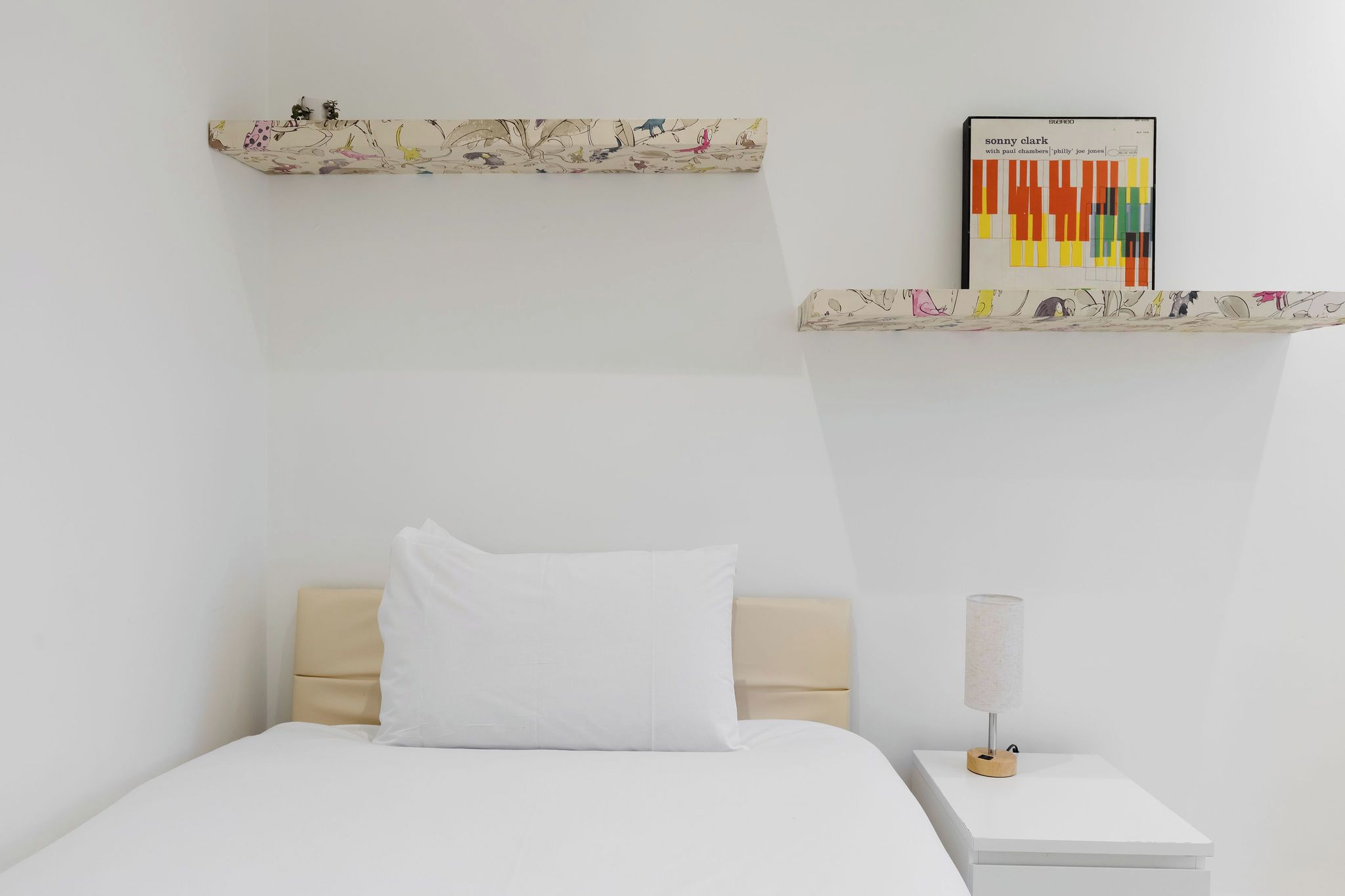 A minimalist hotel room with a bed, a bedside table, a lamp, and two wall-mounted shelves with artwork.