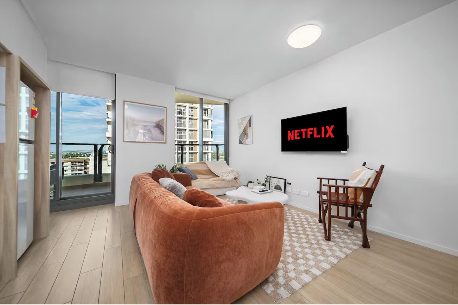Una sala de estar moderna con un sofá grande, una mesa de centro, un televisor montado en la pared y un balcón con vistas a edificios.