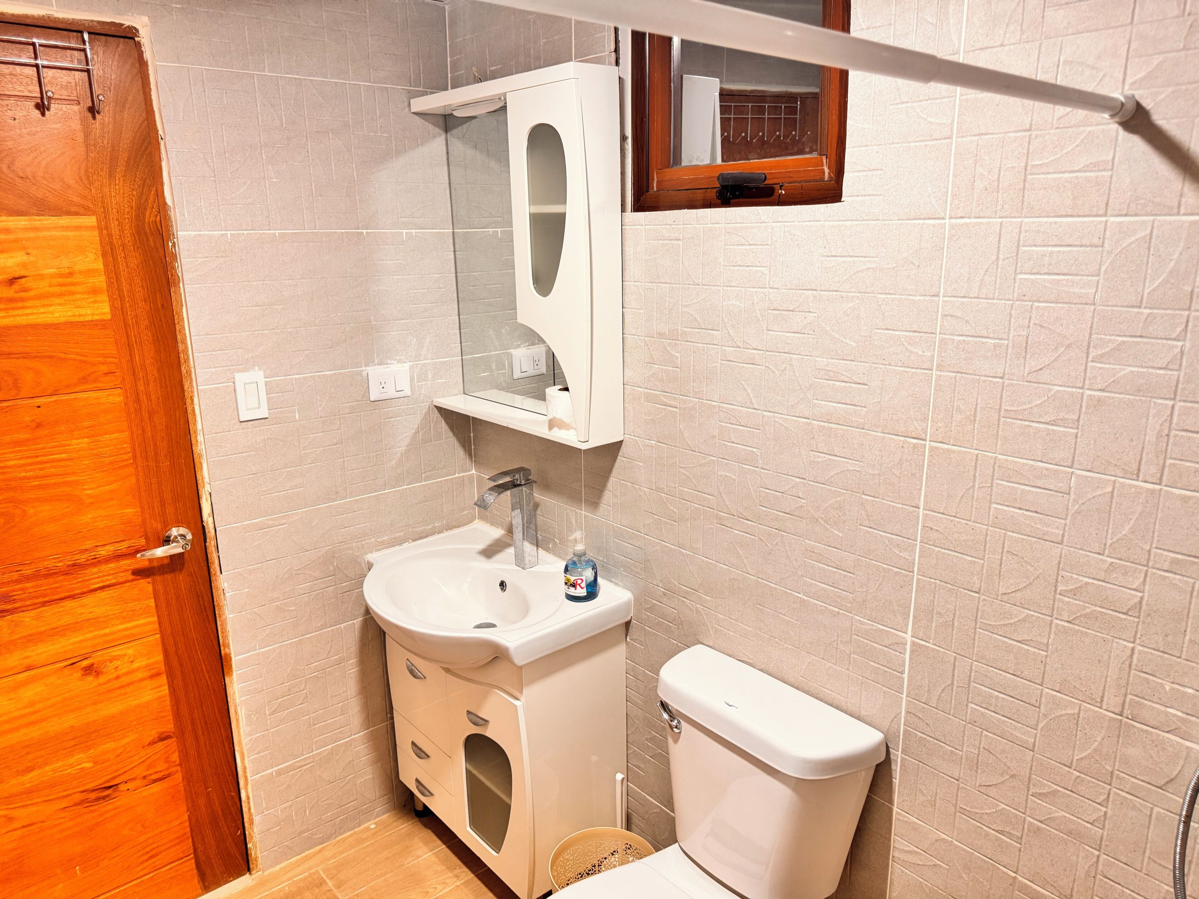 Een badkamer met een houten deur, een wit wastafelmeubel met spiegel, een toilet en een raam met handdoekhouder.