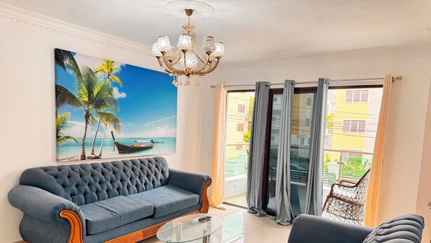 A living room with a blue sofa, a glass coffee table, and a painting of a beach scene. - Santiago Amazing 4bedrooms Apt for Group, Spectacular Wifi, Close to all fun (Santiago de los Caballeros)