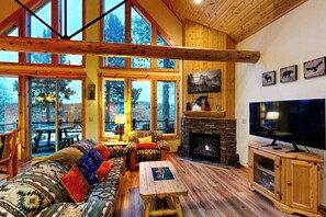 Un vaste salon doté d’un grand plafond en bois, d’une cheminée en pierre, d’un téléviseur et offrant une vue sur l’extérieur grâce à de grandes fenêtres.