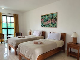A hotel room with two beds, a window with curtains, a painting on the wall, and a small table with a lamp.
