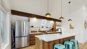 Een moderne keuken met een roestvrijstalen koelkast, houten kastjes, een groot eiland met barkrukken, hanglampen en uitzicht op de stad door een raam.