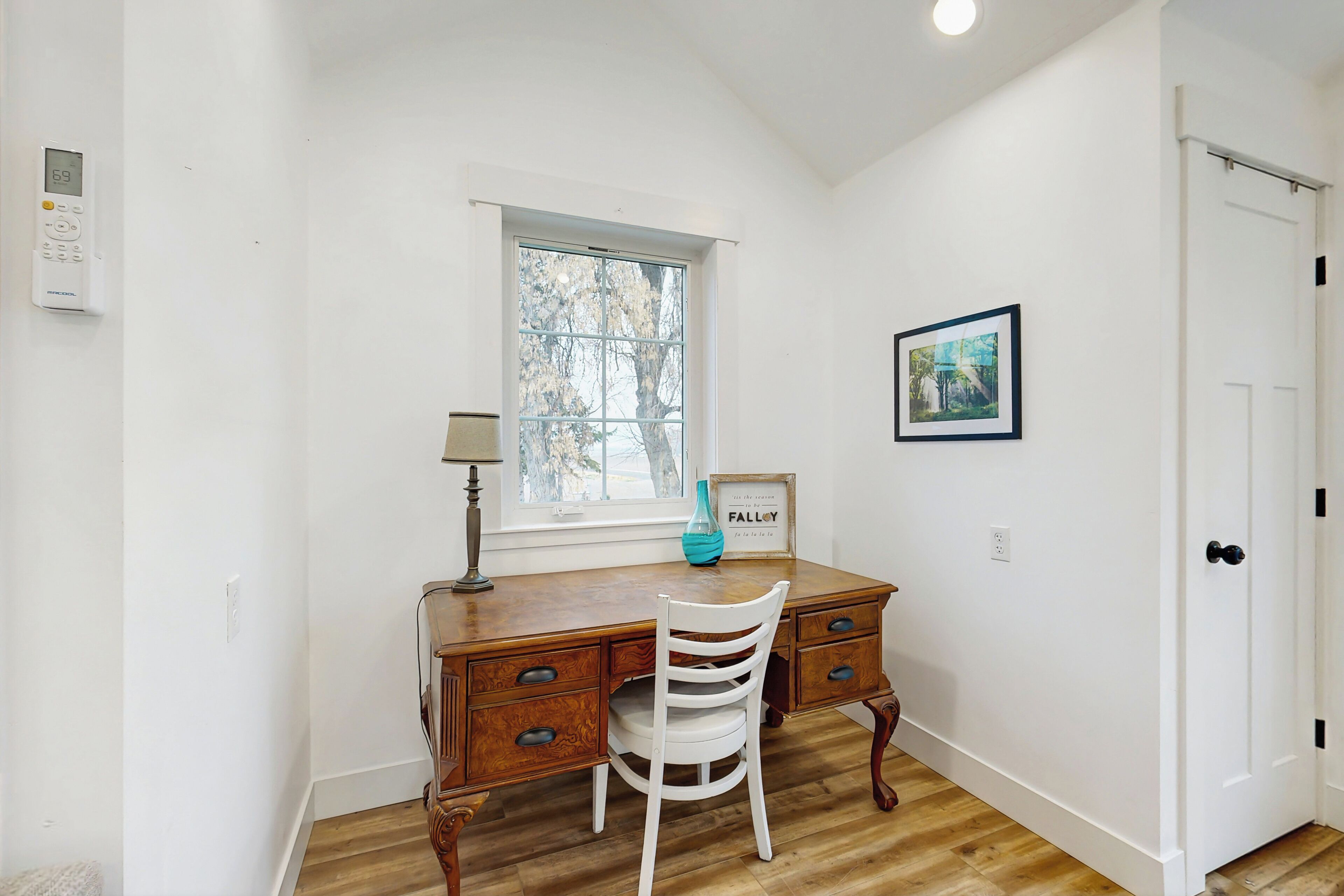 Quarto com escrivaninha de madeira, cadeira branca, janela, quadro na parede e porta.