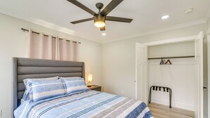 A bedroom with a bed, a ceiling fan, a nightstand, a lamp, a coat rack, and a door leading to another room. - Home 2 Mi to Hermann Park, Zoo & Museum District! (Houston)