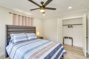 Ein Schlafzimmer mit einem Bett, einem Deckenventilator, einem Nachttisch, einer Lampe, einem Kleiderständer und einer Tür zu einem anderen Raum.