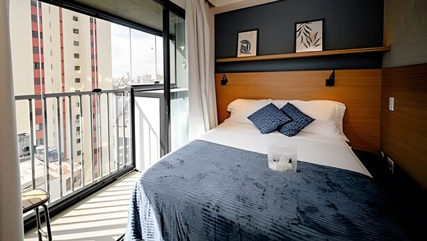A hotel room with a large window, a bed with pillows, a bedside table, and a wooden headboard. - UENO 906 (Sao Paulo)