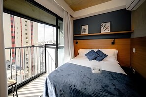 Habitación de hotel con una ventana grande, una cama con almohadas, una mesita de noche y un cabecero de madera.
