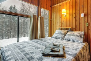 Une chambre avec une grande fenêtre, des murs en bois, un lit recouvert d’une courtepointe à motifs, deux oreillers et une lampe de chevet.