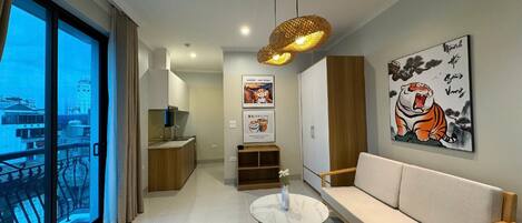 A modern living room with a sofa, a round coffee table, and a large window with a view of buildings. There is a kitchen area with cabinets and a hanging light fixture.