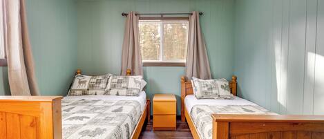A bedroom with two wooden beds, a window with curtains, a ceiling fan, and a small wooden nightstand between the beds.