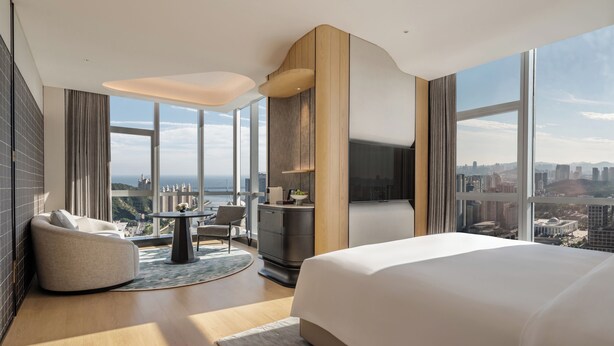 A modern hotel room with a large bed, a round table, a sofa, and a view of the cityscape. - Pan Pacific Dalian (Dalian)