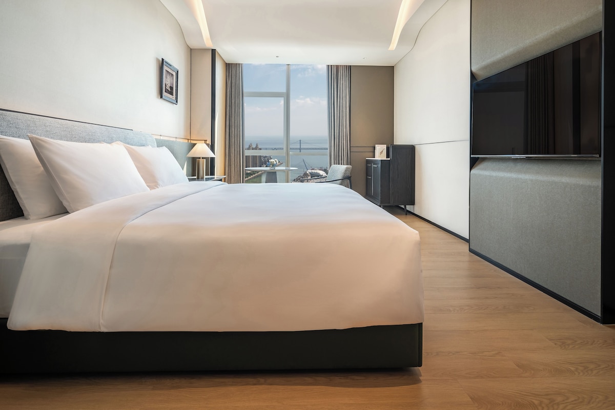 A modern hotel room with a large bed, a desk, a chair, and a TV.