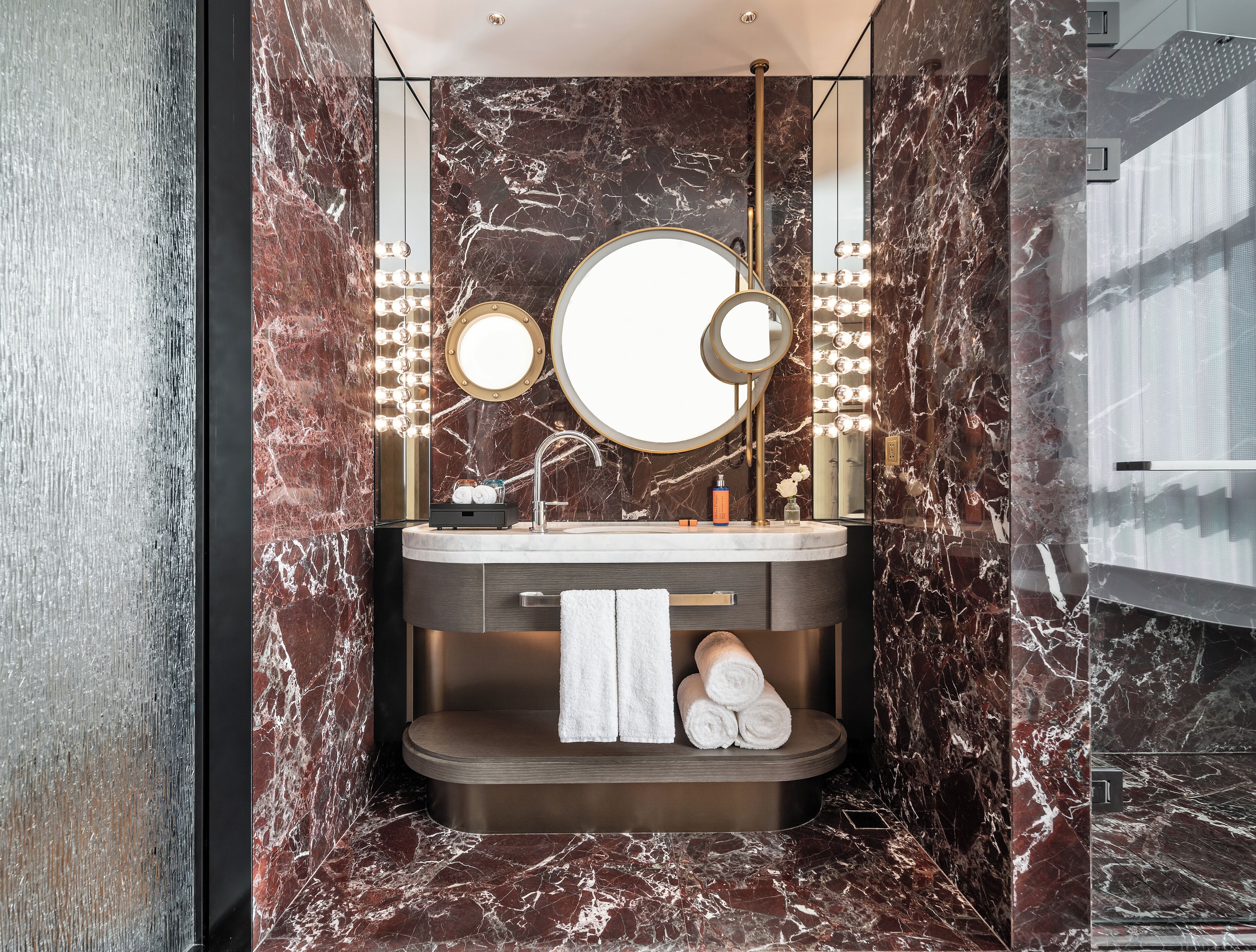 A modern bathroom with marble walls, a vanity with a round mirror, and a built-in shelf with towels.