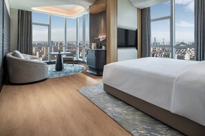 A modern hotel room with a large bed, a curved seating area, a small table, and a city view through large windows.