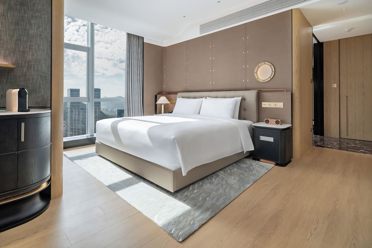 Ein modernes Hotelzimmer mit einem großen Bett, einem Nachttisch, einer Lampe und Blick auf die Stadt.