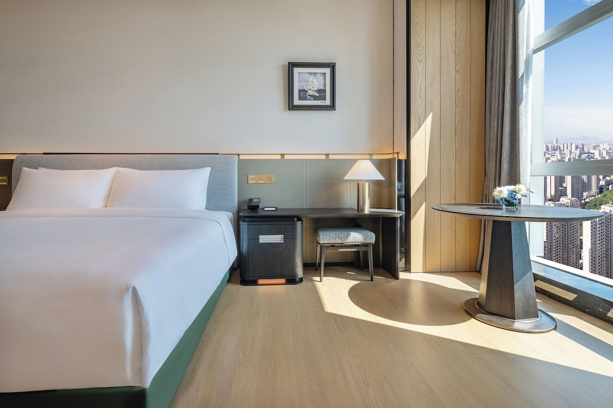 A hotel room with a bed, desk, chair, and a small table with a lamp.