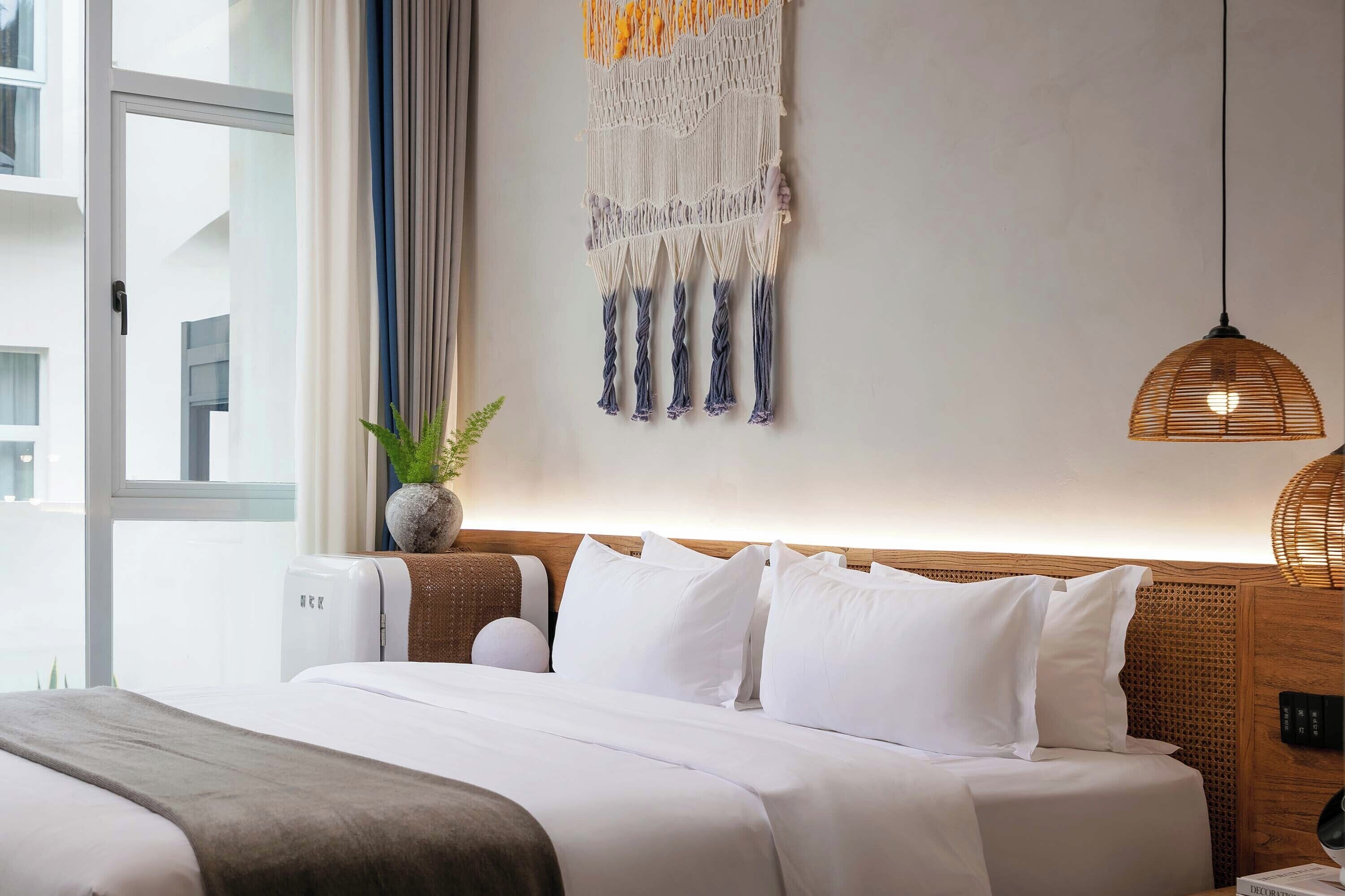 A hotel room with a bed, a wall-mounted macrame piece, a potted plant, and a hanging light fixture.